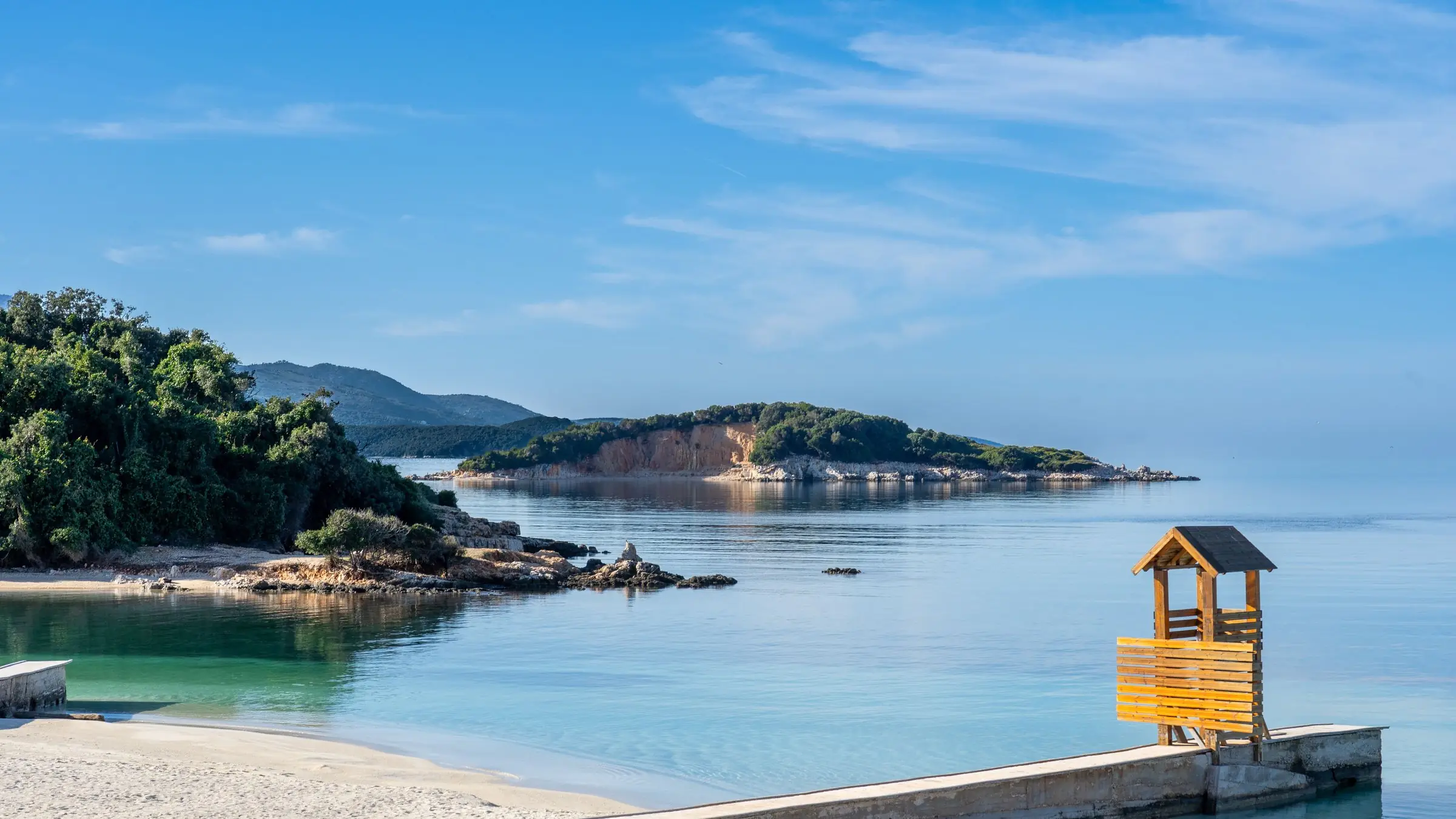Plajă cu nisip alb și insulă împădurită în apa limpede din Ksamil, Albania